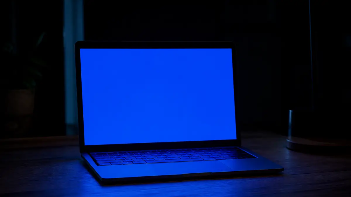 Open laptop in a dark room filling the wooden desk with a solid blue glow, no letters or logos