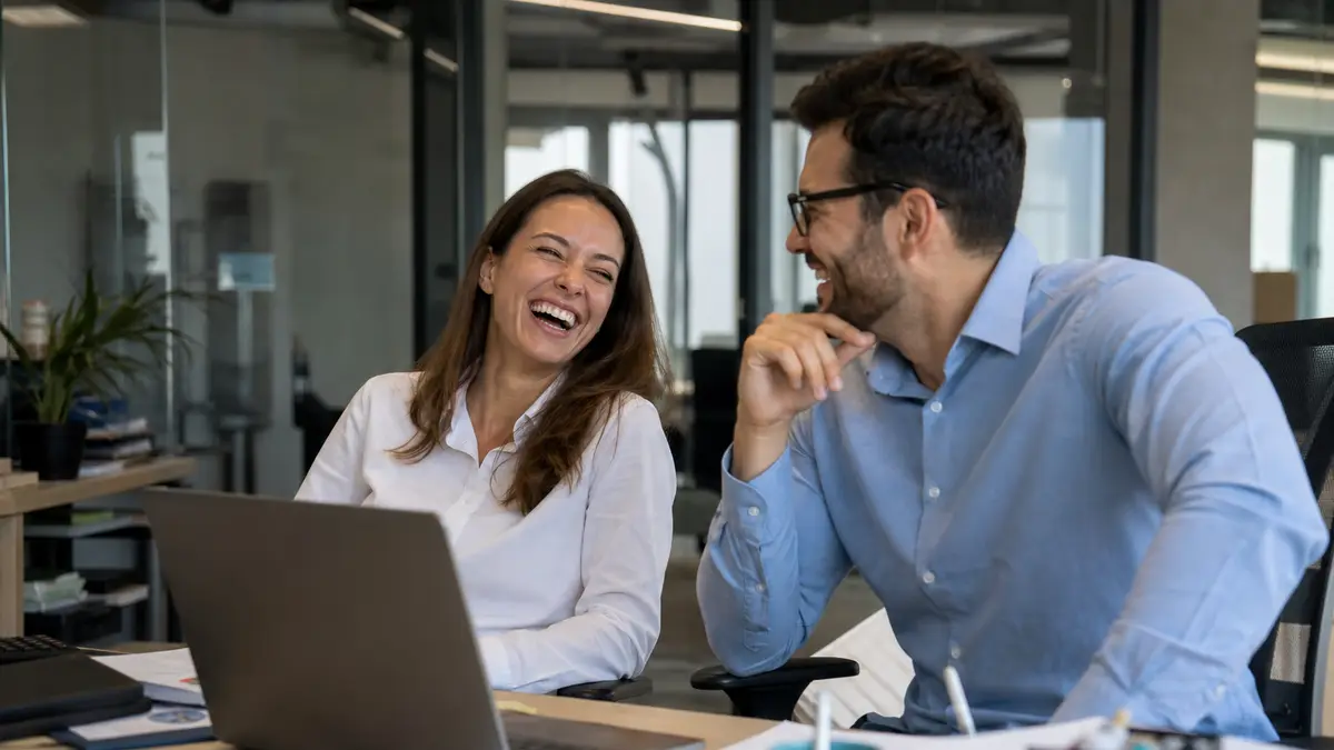 Two coworkers laughing beside a messy desk after a short joke, laptop screen not readable
