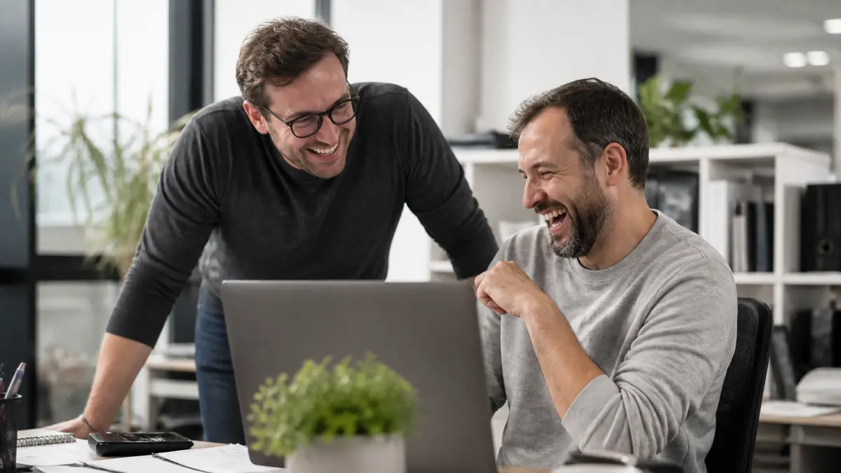 Two coworkers laughing beside a messy desk after a short joke, laptop turned away