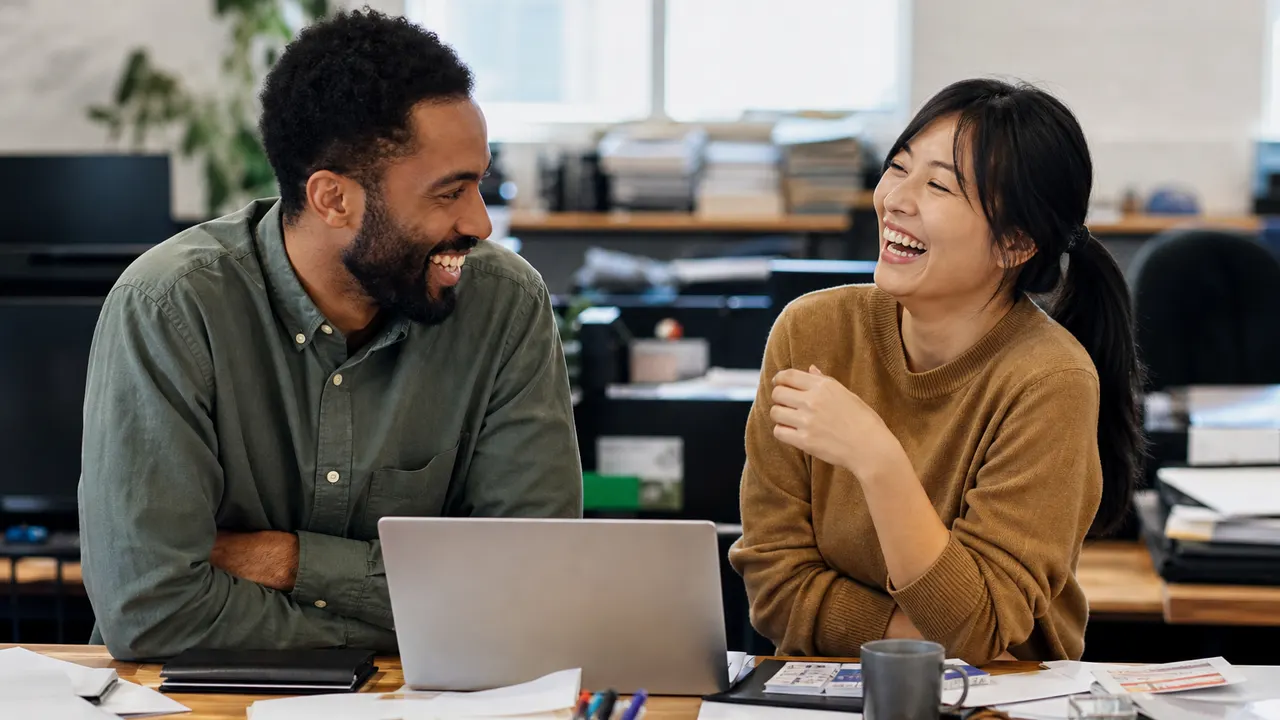 Two coworkers laughing beside a messy desk after a short joke, laptop screen not readable