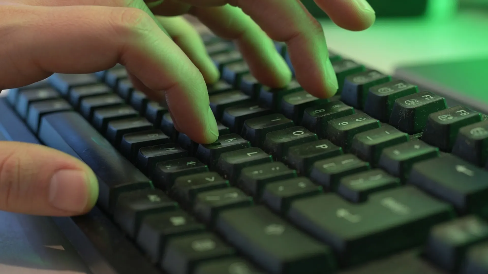 Close-up of fingers pressing keys on a dark keyboard under green monitor light, no on-screen letters